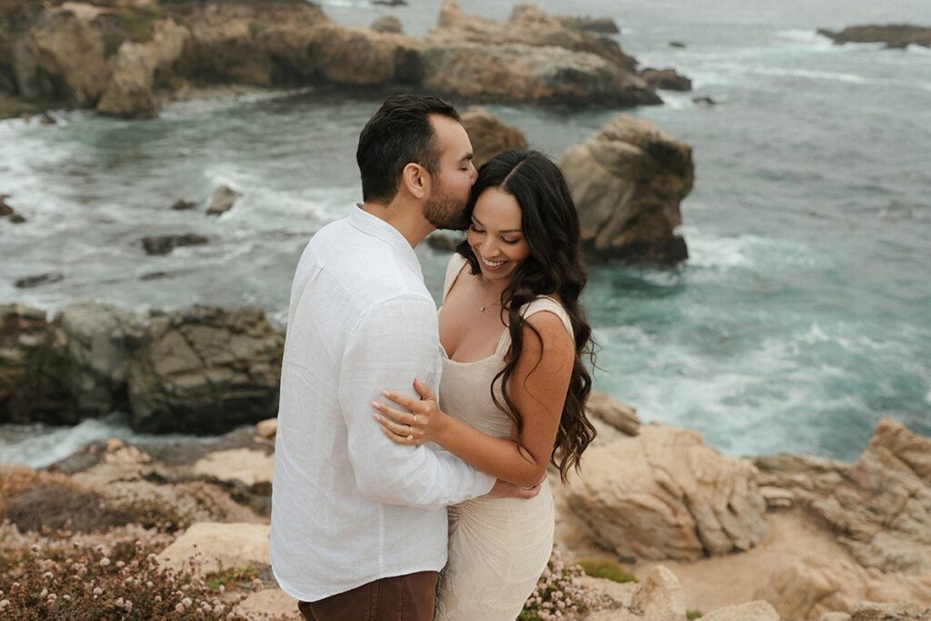 A man kissing a woman on the forehead while embracing on a rocky cliff overlooking the ocean during an engagement session near Rose Garden San Jose