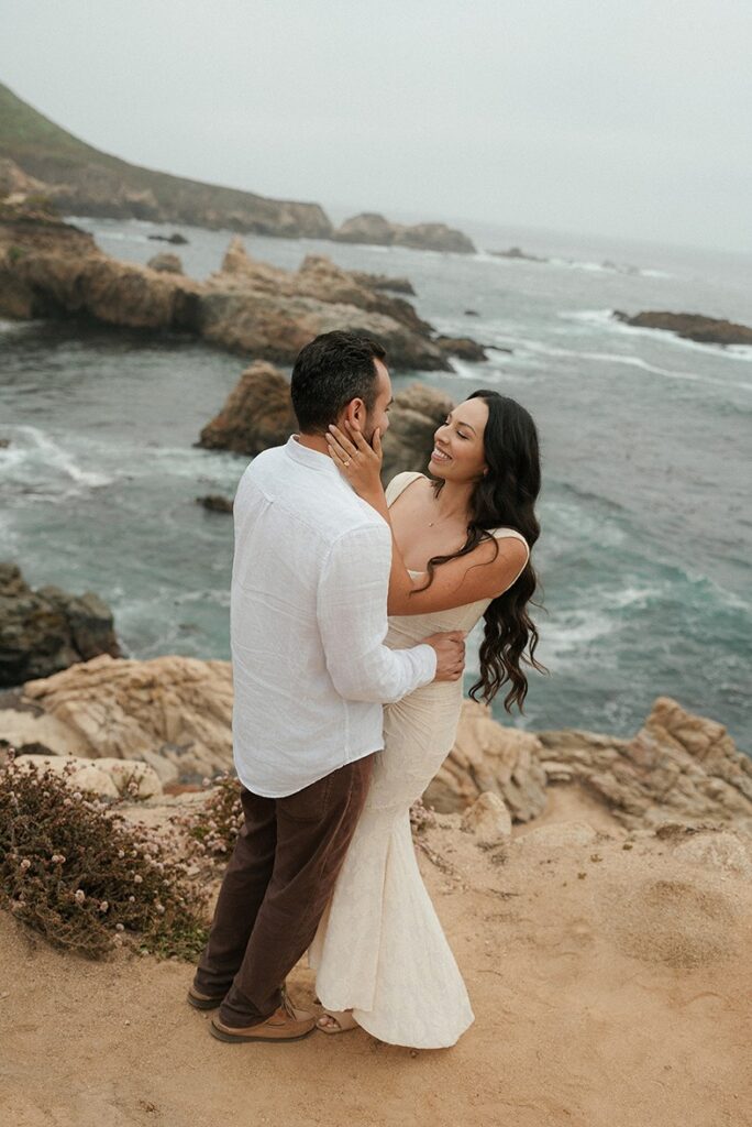 A man and woman embracing on a high rocky cliff overlooking a dramatic turquoise ocean with crashing waves along the Santa Cruz coastline.