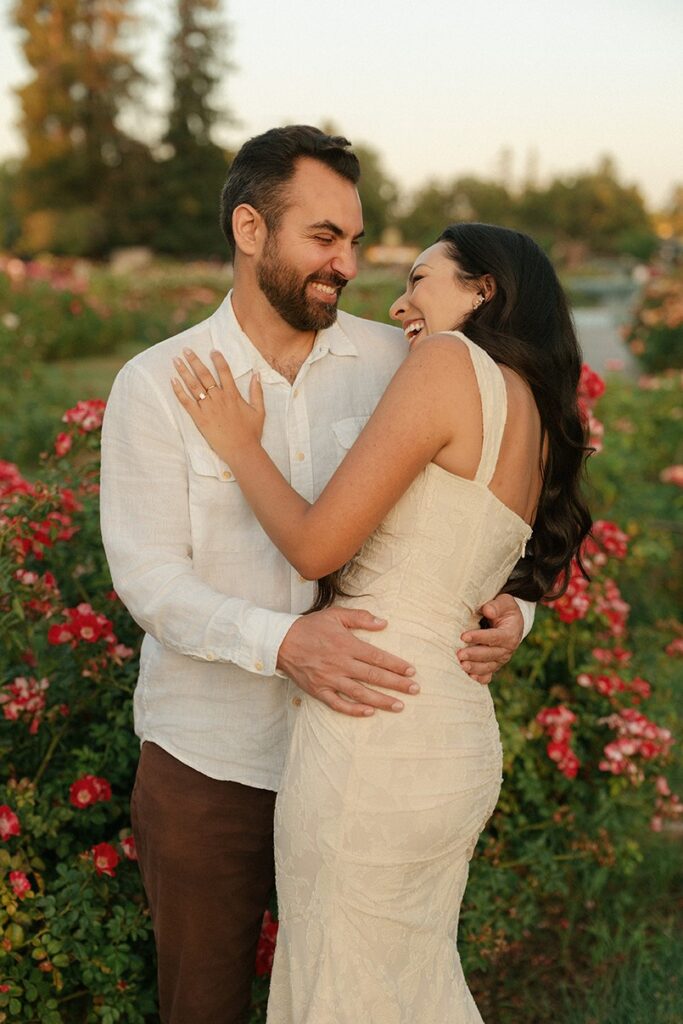 An engaged couple sharing a laugh together while standing among vibrant red rose bushes in San Jose.
