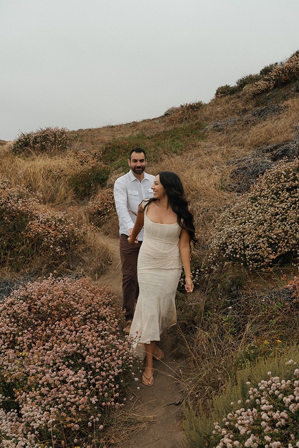 An engaged couple walking along a scenic dirt path on a grassy coastal bluff during an outdoor portrait session near Santa Cruz