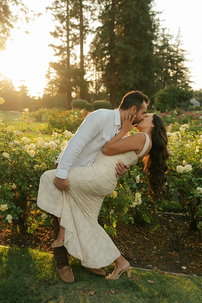A man dipping and kissing his partner on a grass lawn surrounded by blooming white roses during a golden hour engagement session.