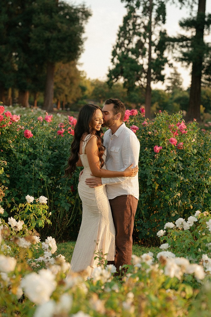 A man kissing a woman on the cheek while standing along a pathway lined with red and white rose bushes in San Jose.