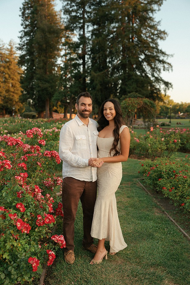 An engaged couple holding hands and smiling together while standing on a grassy path next to vibrant rose garden flower beds.