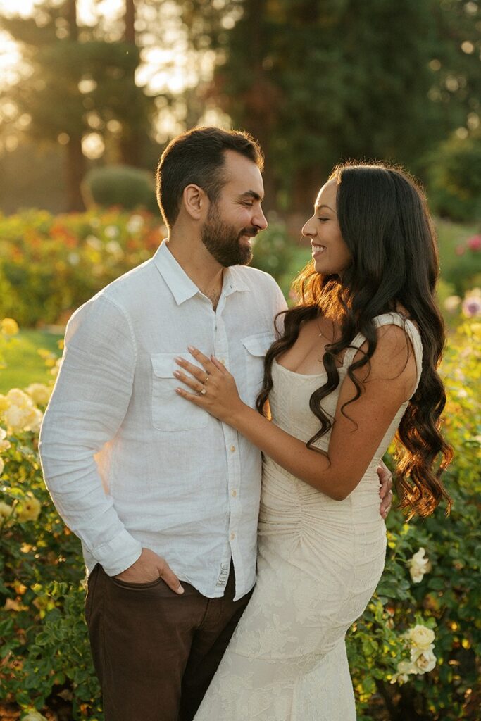 A man and woman smiling at each other while embracing in front of blooming rose bushes during a golden hour engagement session at the San Jose Municipal Rose Garden.