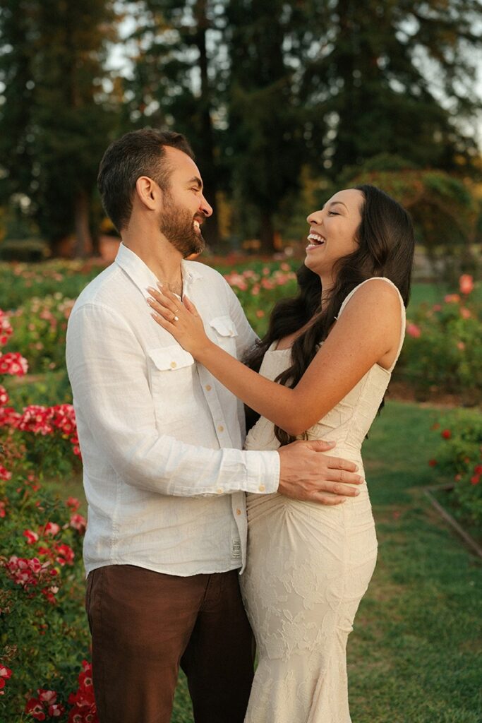 A woman laughing joyfully while holding her partner's shirt collar during a romantic couple's portrait session at a San Jose rose garden.