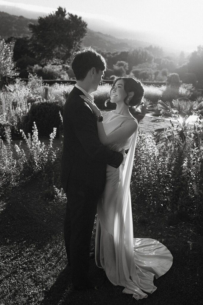 A black and white photograph of a bride and groom embracing lovingly amidst tall ornamental grasses in a sunlit garden at Filoli.