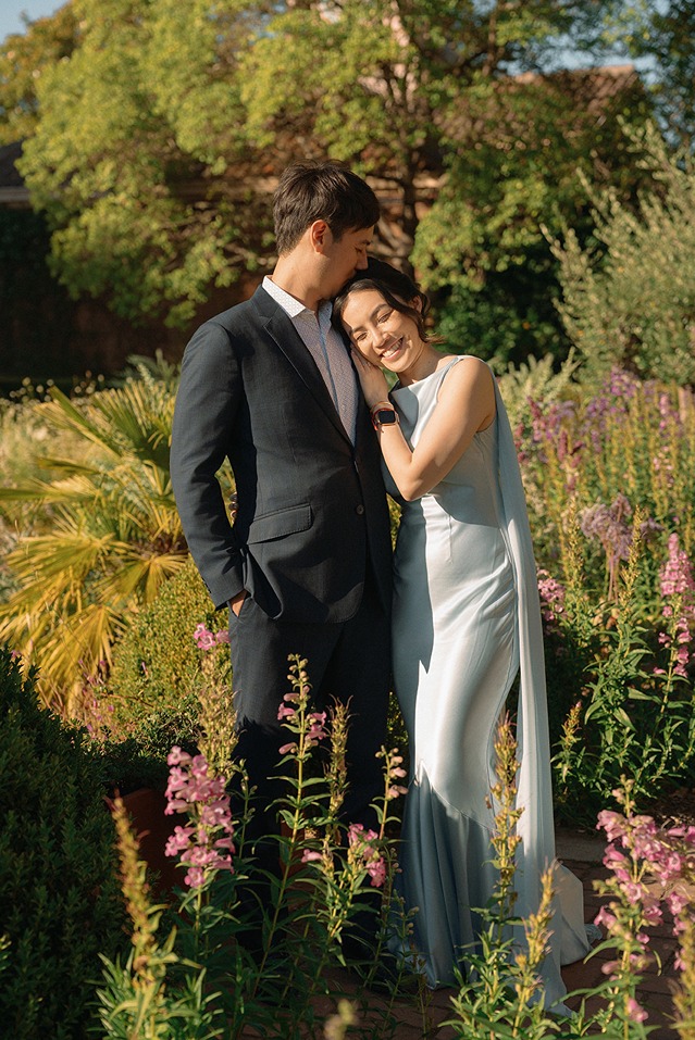 A bride leans her head gently on the groom's shoulder while standing amidst lush flowering plants and golden-lit foliage.