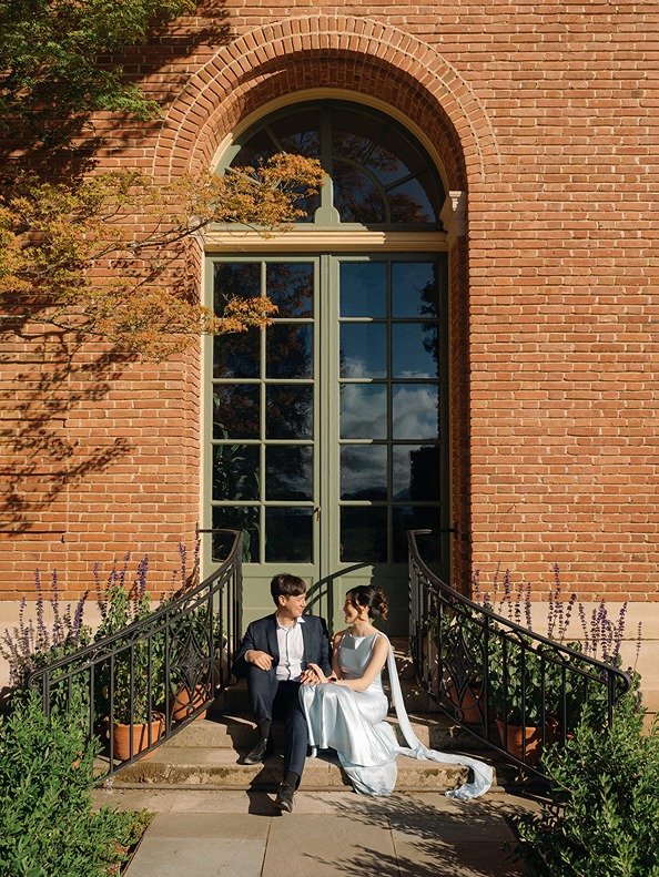 A newlywed couple sits on the stone steps beneath a grand arched glass window of the brick estate building, bathed in soft afternoon light.