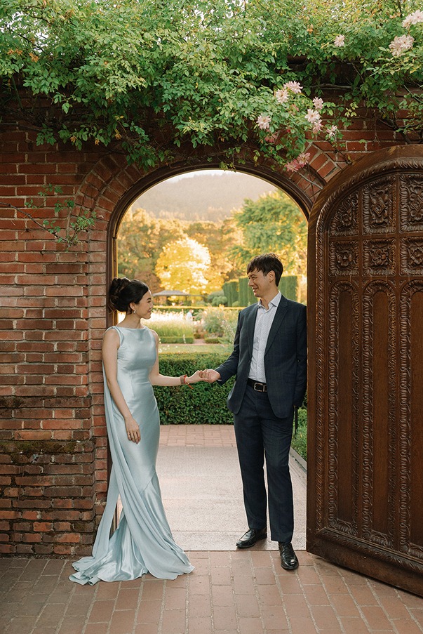 A wedding couple poses inside a historic brick archway with a heavy wooden door at Filoli, looking out toward a sun-drenched garden courtyard.