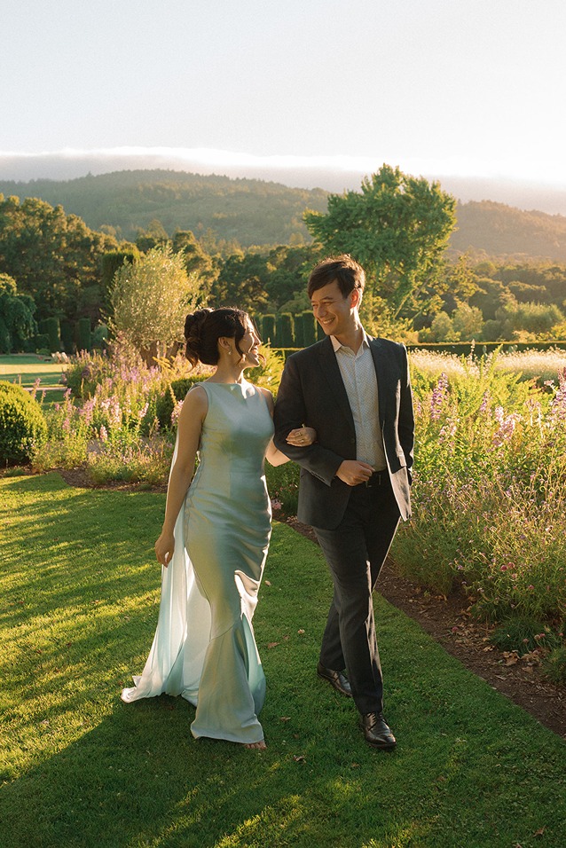 A bride in a sleek white gown and a groom in a dark suit walk hand-in-hand across a grassy field at Filoli during golden hour, with soft hills visible in the background.
