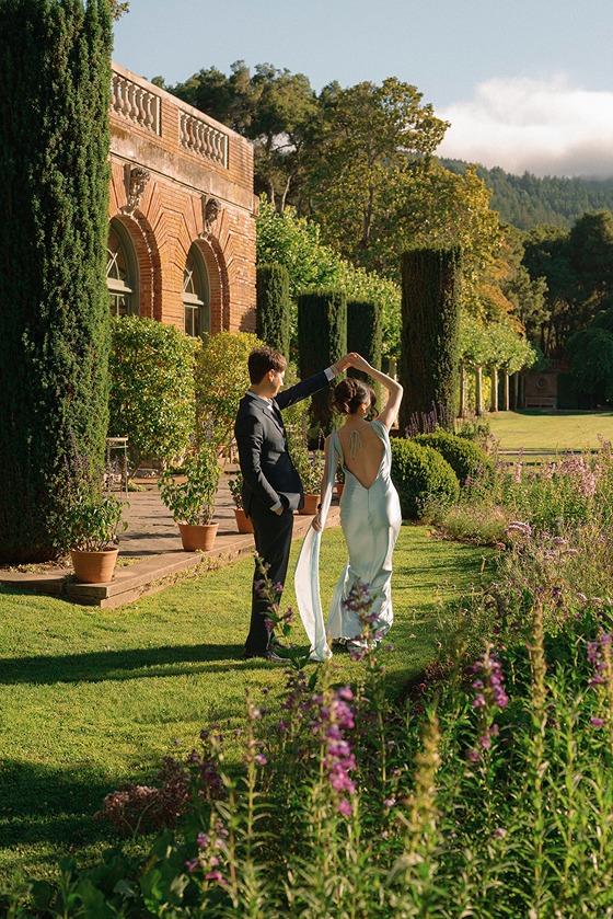 A bride and groom stand together on a wide green lawn next to a grand brick estate building, bordered by tall cypress trees and wild garden blooms.