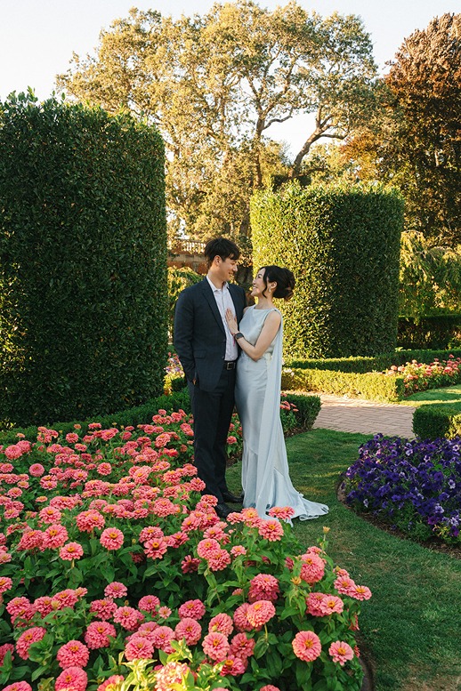 A wedding couple stands together next to a vibrant bed of pink flowers at Filoli, backed by tall, perfectly manicured green hedges and sunny trees.