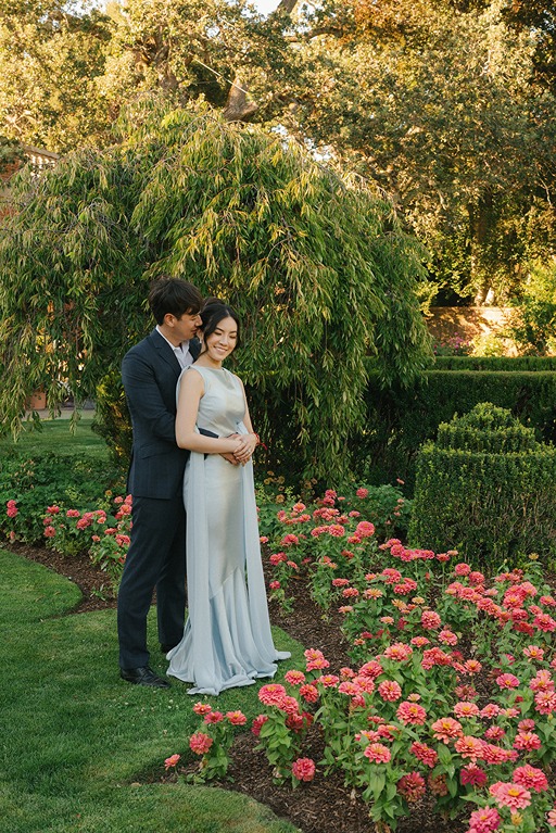 A wedding couple stands lovingly on a manicured lawn next to a vibrant bed of pink flowers, framed by structured hedges and weeping trees.