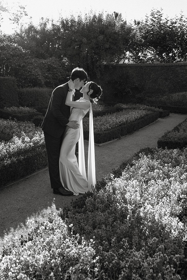 A black and white photograph of a bride and groom kissing on a walkway in the structured formal gardens, surrounded by low hedges and sun-dappled flowerbeds.
