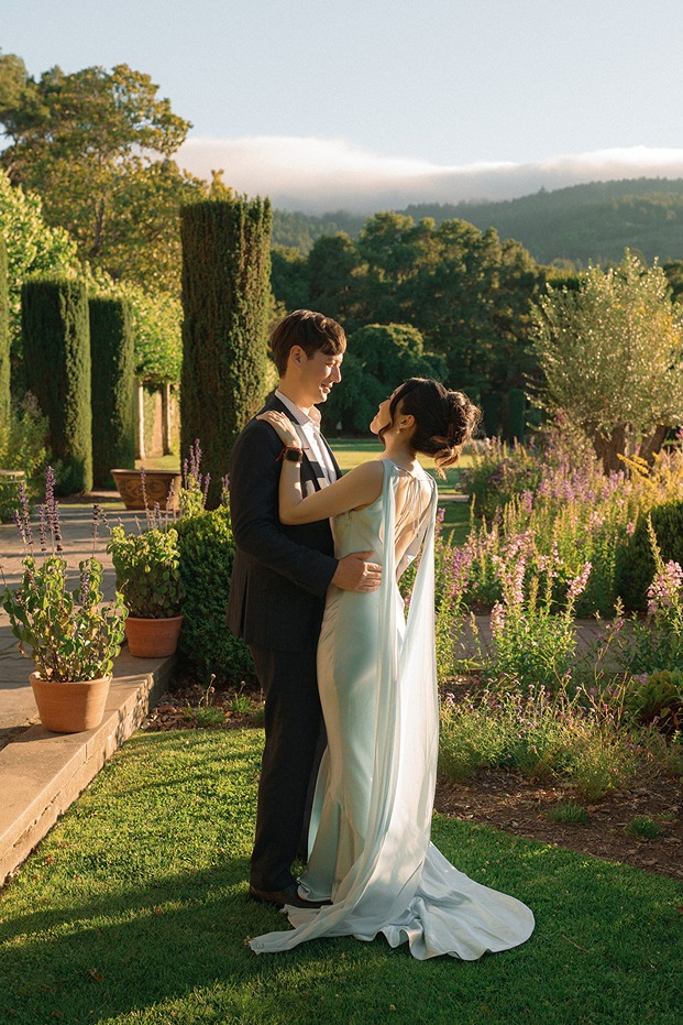A wedding couple embraces on a lush green lawn during a portrait session at Filoli Historic House & Garden, featuring towering yew trees and a vibrant English Renaissance-style garden backdrop bathed in warm afternoon sunlight.