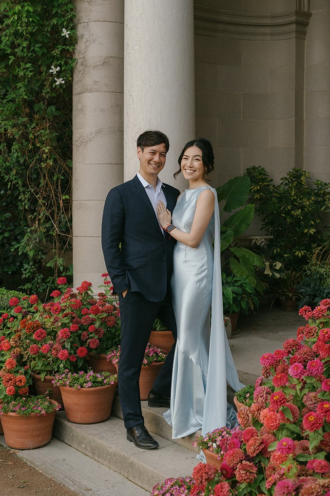 A newlywed couple stands together smiling next to a large stone pillar at Filoli, surrounded by potted pink flowering plants and lush green foliage.