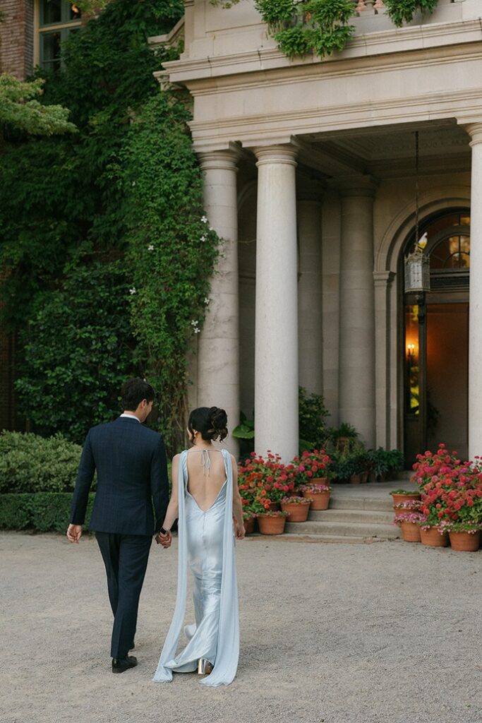 A bride and groom walk away from the camera toward the historic stone columned entrance of the Filoli mansion, framed by ivy-covered walls and potted flowers.