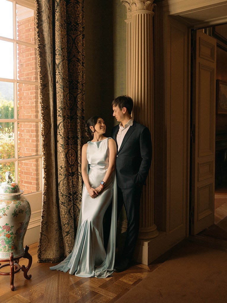 A wedding couple stands indoors by a large window inside the historic Filoli mansion, illuminated by soft natural light filtering past heavy drapery next to an ornate pillar.
