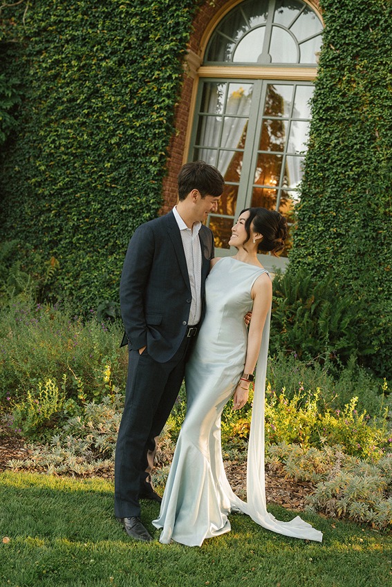 A wedding couple shares an intimate moment standing against an ivy-covered brick wall next to a large arched window .