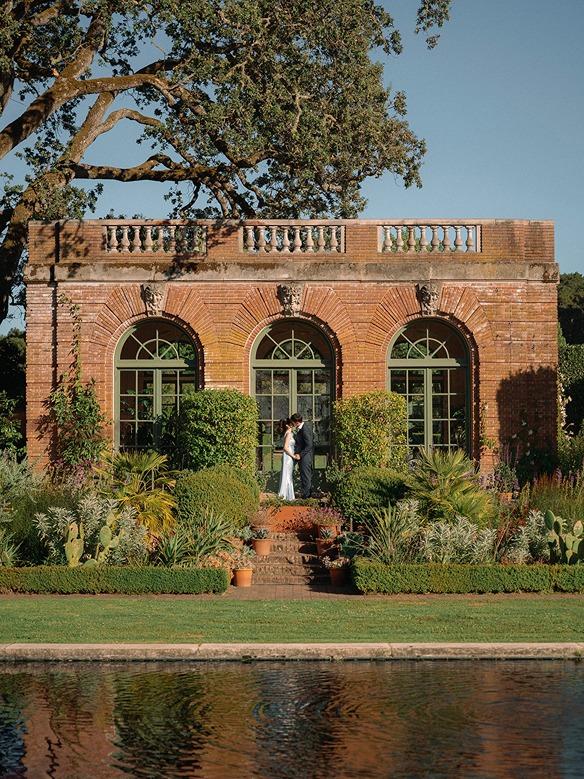 The historic brick Garden House structure at Filoli sits behind a calm reflecting pool, featuring three large arched glass doors, climbing green ivy, and a stone balustrade roof line against a clear blue sky.