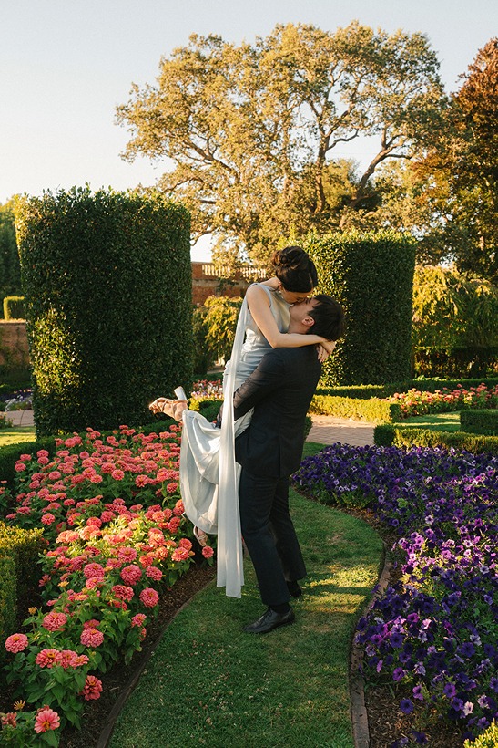 A groom lifts and spins his bride along a garden path, surrounded by colorful flowerbeds, manicured hedges, and sunlit oak trees.