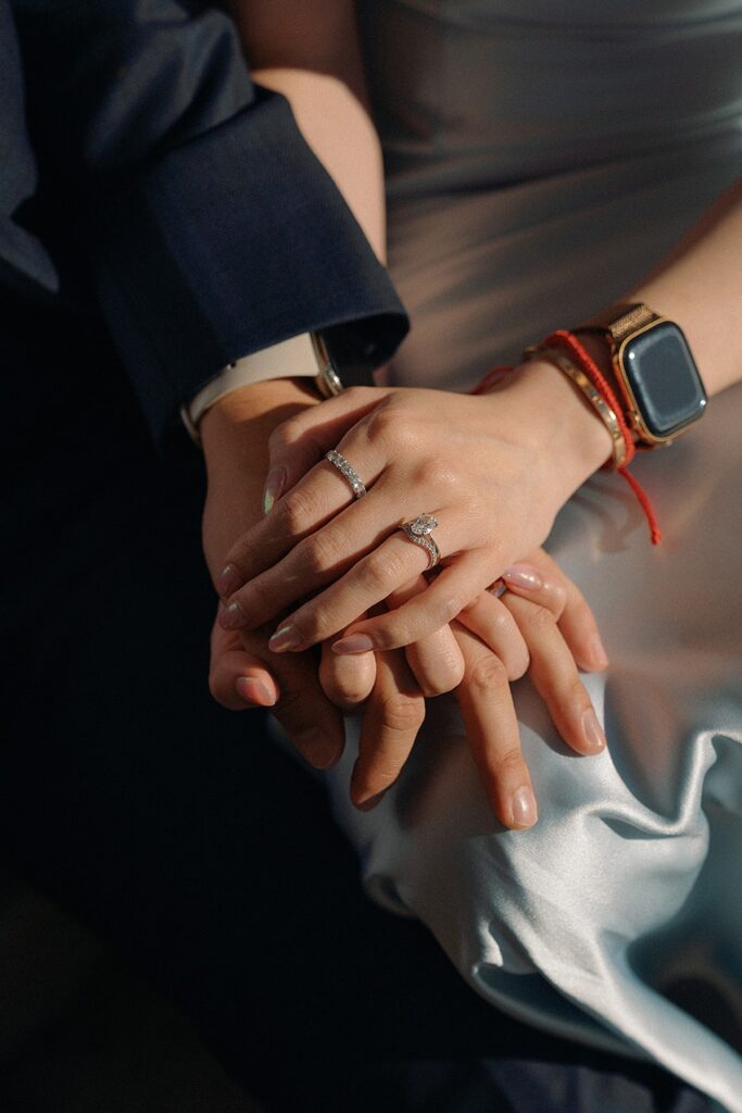 A close-up shot of a bride and groom's intertwined hands resting on silk wedding gown fabric ,showcasing their wedding rings and a watch under dramatic soft lighting.