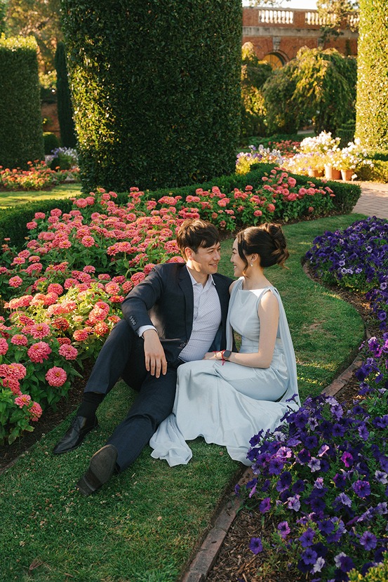 A wedding couple sits together on a lush green lawn, surrounded by curved flowerbeds filled with vibrant pink and purple blossoms and high-cut formal hedges.