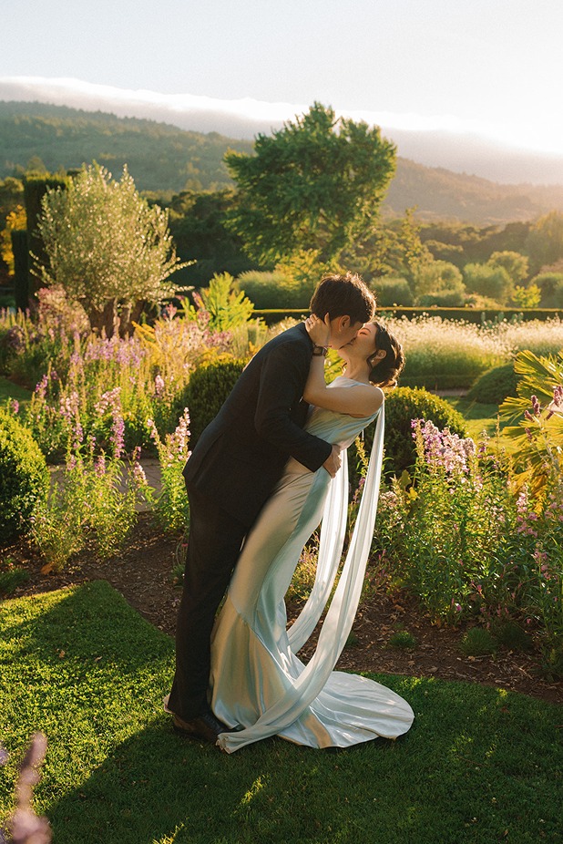 A groom dips and kisses his bride on a grassy lawn at Filoli during golden hour, surrounded by wild garden grasses and misty sunlit hills in the background.